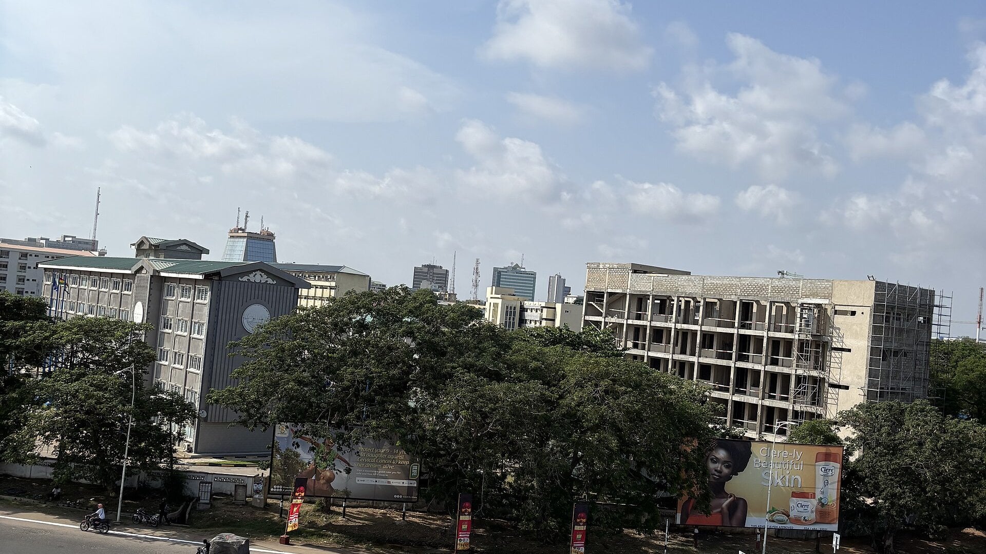 Government and office buildings with trees and an unfinished block under construction in central Accra, Ghana, under a partly cloudy sky