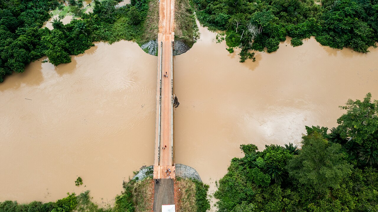 Aerial view of a bridge crossing a wide muddy brown river surrounded by green forest