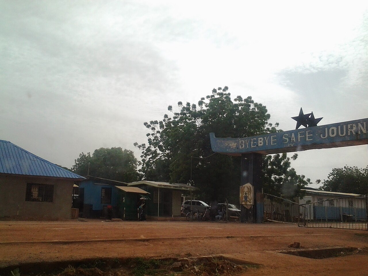 A blue arch topped with two black stars reading Bye Bye Safe Journey spans a dirt road at Ghana's border post with Burkina Faso
