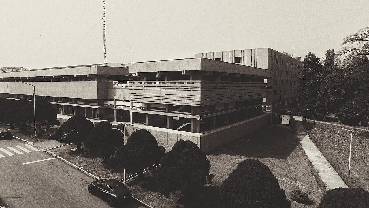 A large concrete university building with balconies and trimmed trees beside a campus road, in sepia tones