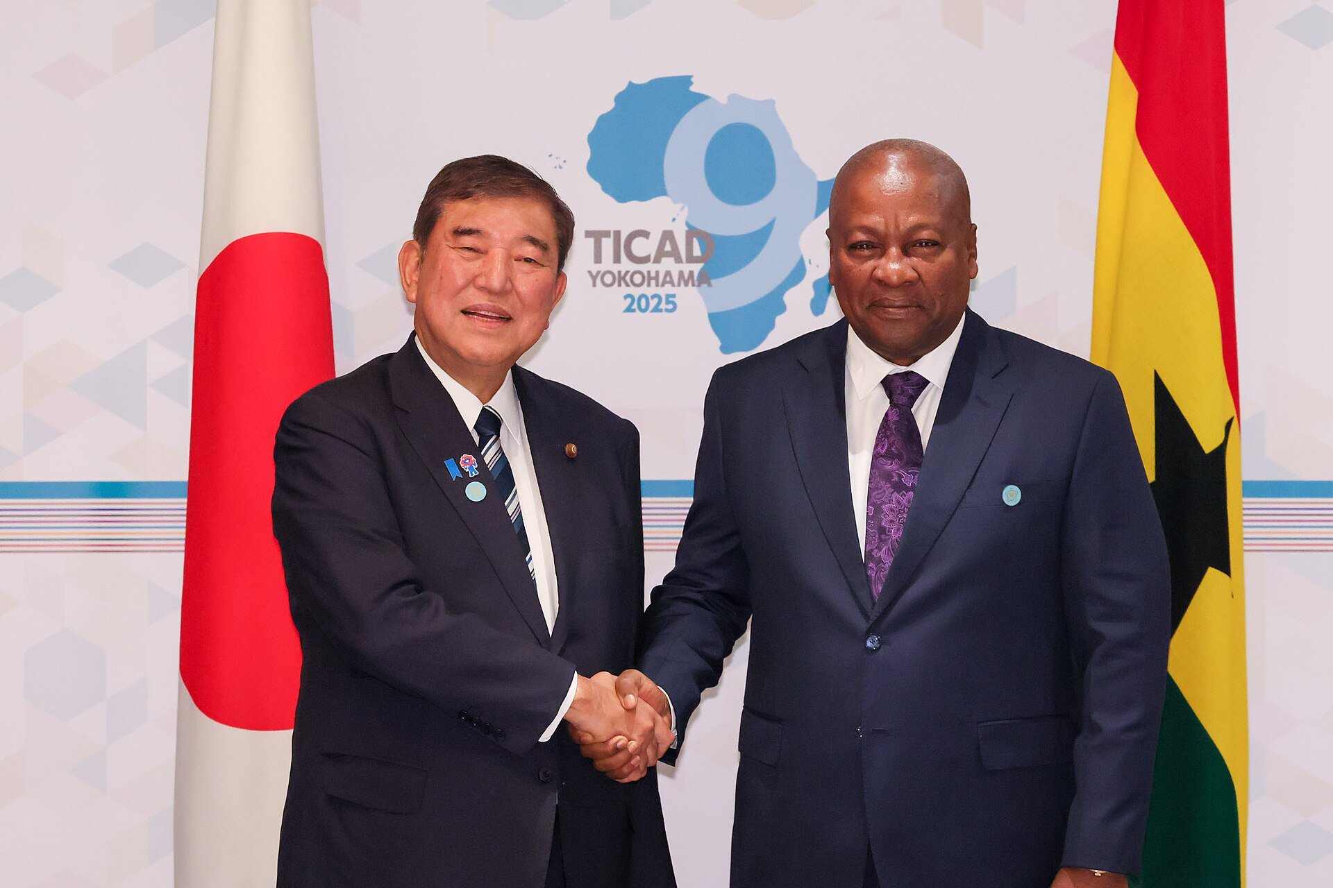 President John Dramani Mahama of Ghana shaking hands with the prime minister of Japan in front of the Ghana and Japan flags at the TICAD 9 conference