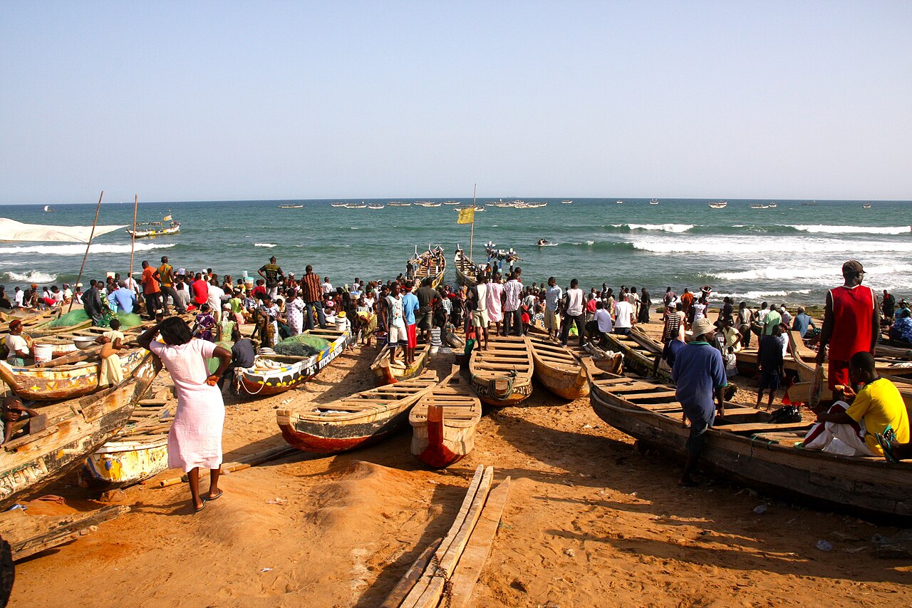 Crowds and painted wooden fishing canoes on the sand at Prampram beach, Ghana, with more boats out at sea