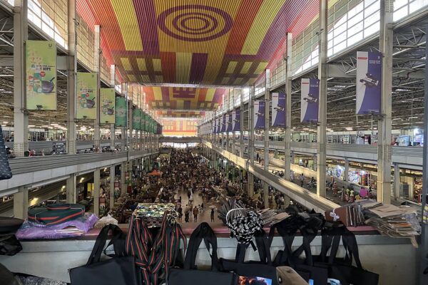 Crowded two-storey market hall in Kumasi, Ghana, with shoppers in the central aisle under a colourful striped ceiling