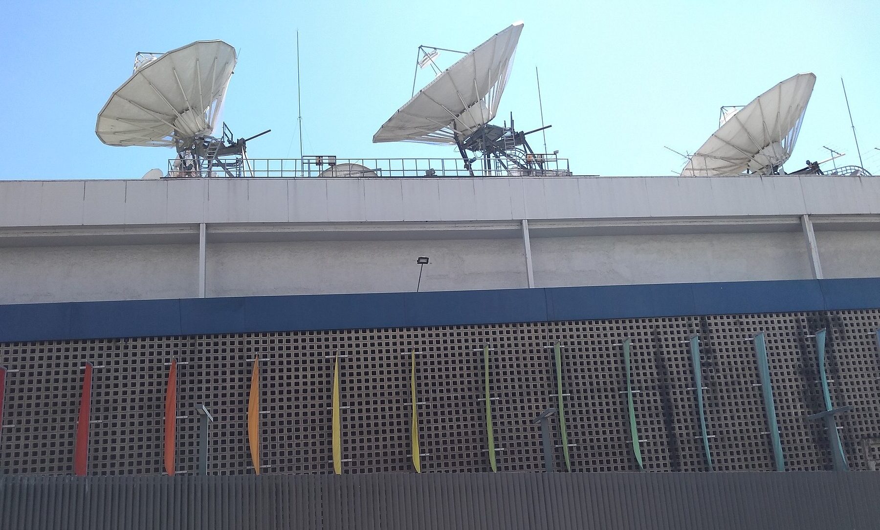 Three satellite dishes on the roof of the TV Globo headquarters in Rio de Janeiro above a wall of lattice blocks hung with coloured banners
