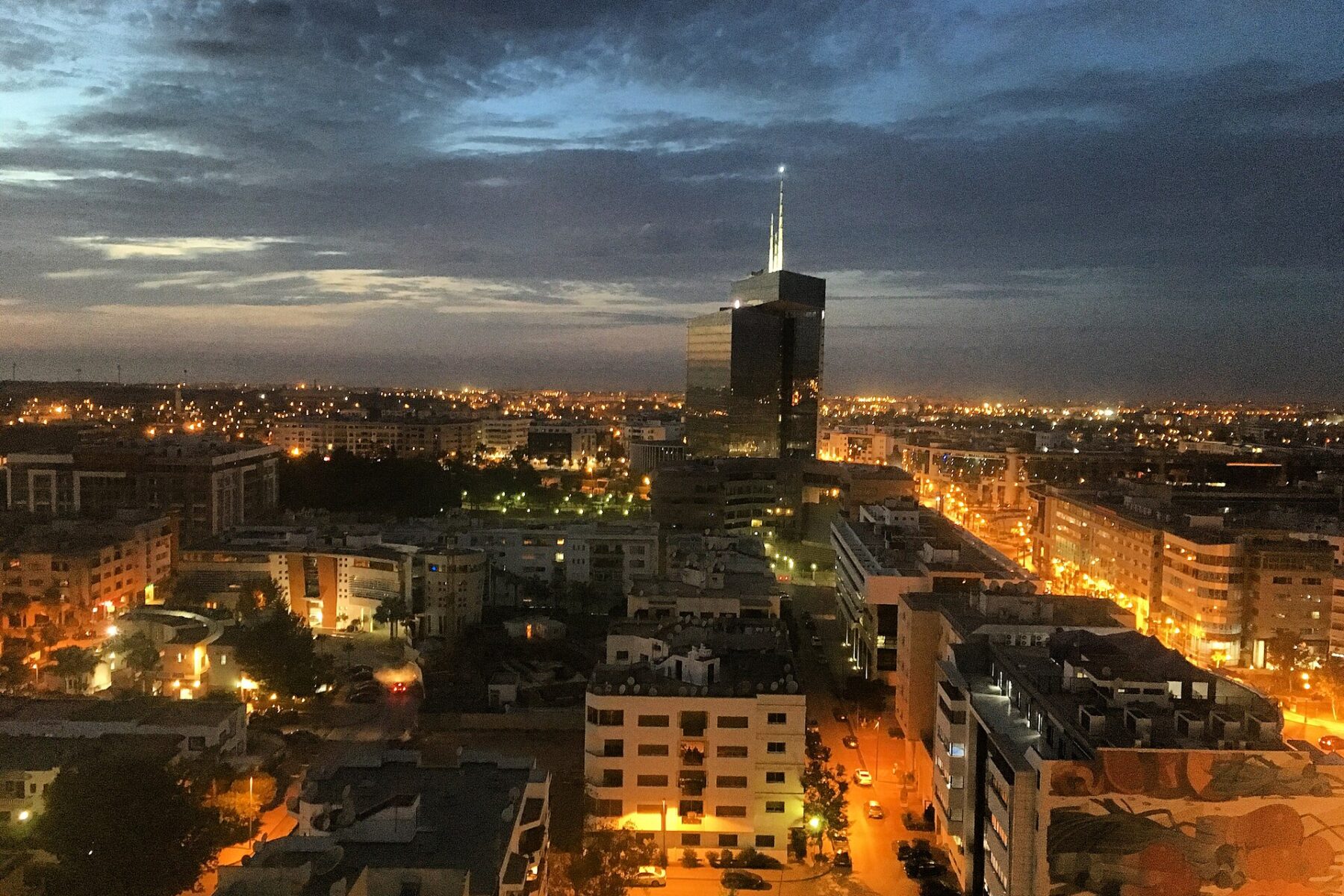 Rabat city lights at dusk under a stormy evening sky, with a glass office tower rising above apartment blocks