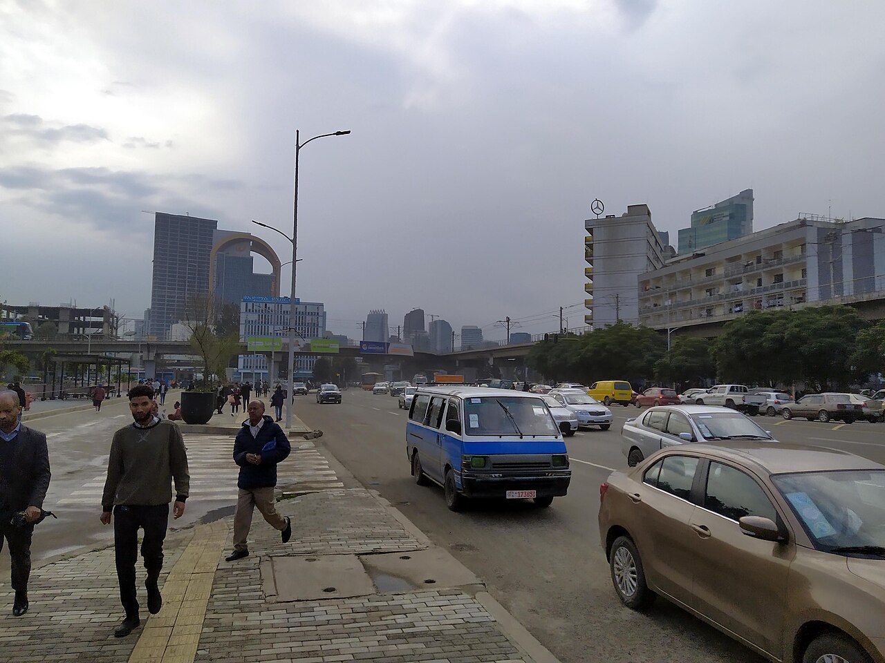 Pedestrians and a blue-and-white minibus taxi on a wide road at Meskel Square in Addis Ababa, with high-rise buildings under a cloudy sky
