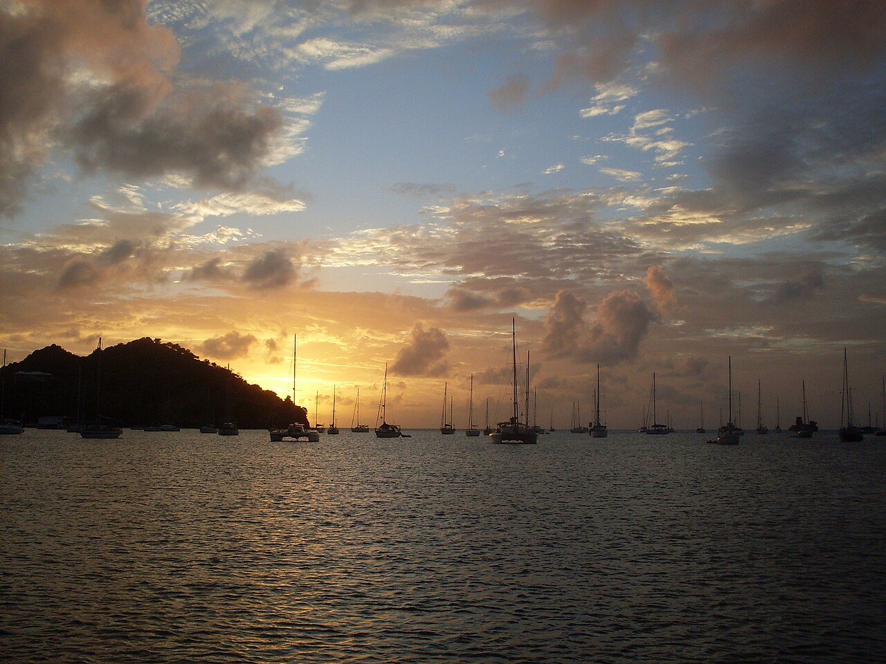 Sailing boats at anchor in a bay at sunset, with a dark hill on the left and orange clouds over the sea.