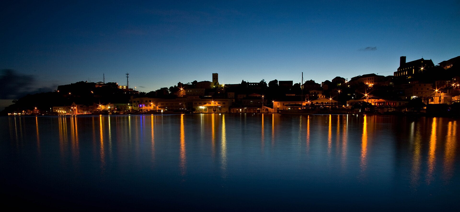 The lit-up waterfront of St George's Carenage harbour at dusk, reflected in calm dark water under a deep blue sky.