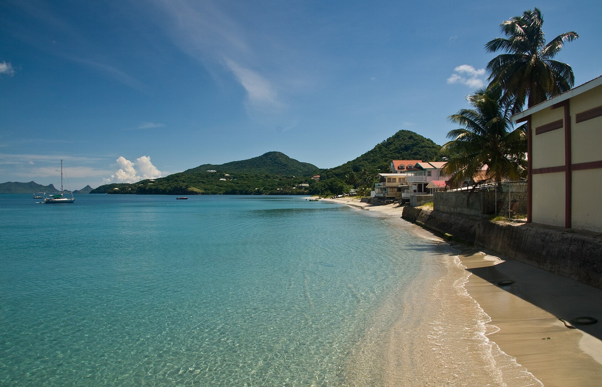 Clear turquoise water and a sandy shore at Hillsborough on Carriacou, with green hills, a few houses and a sailing boat at anchor.