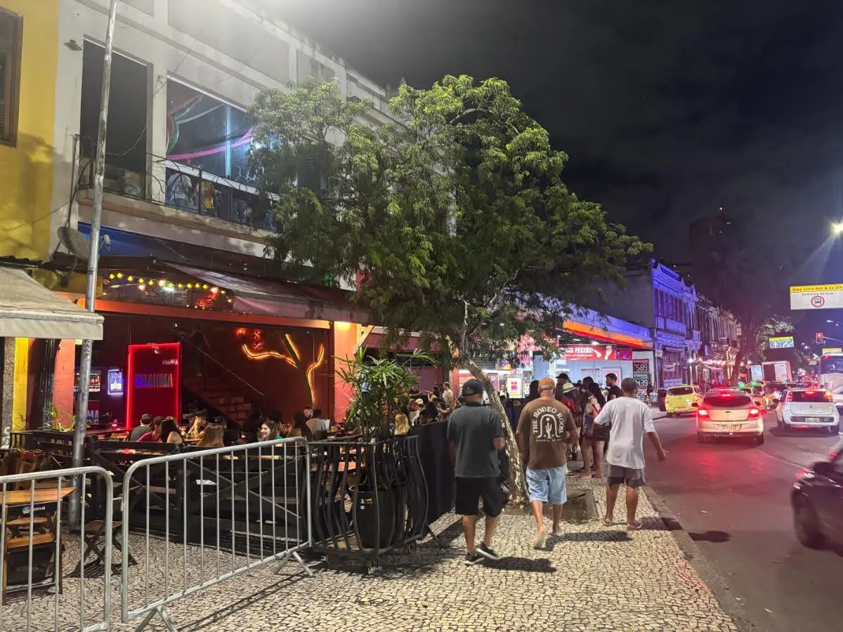 A busy Lapa street at night in Rio de Janeiro, with outdoor bar tables, cobbled pavement and passing cars.