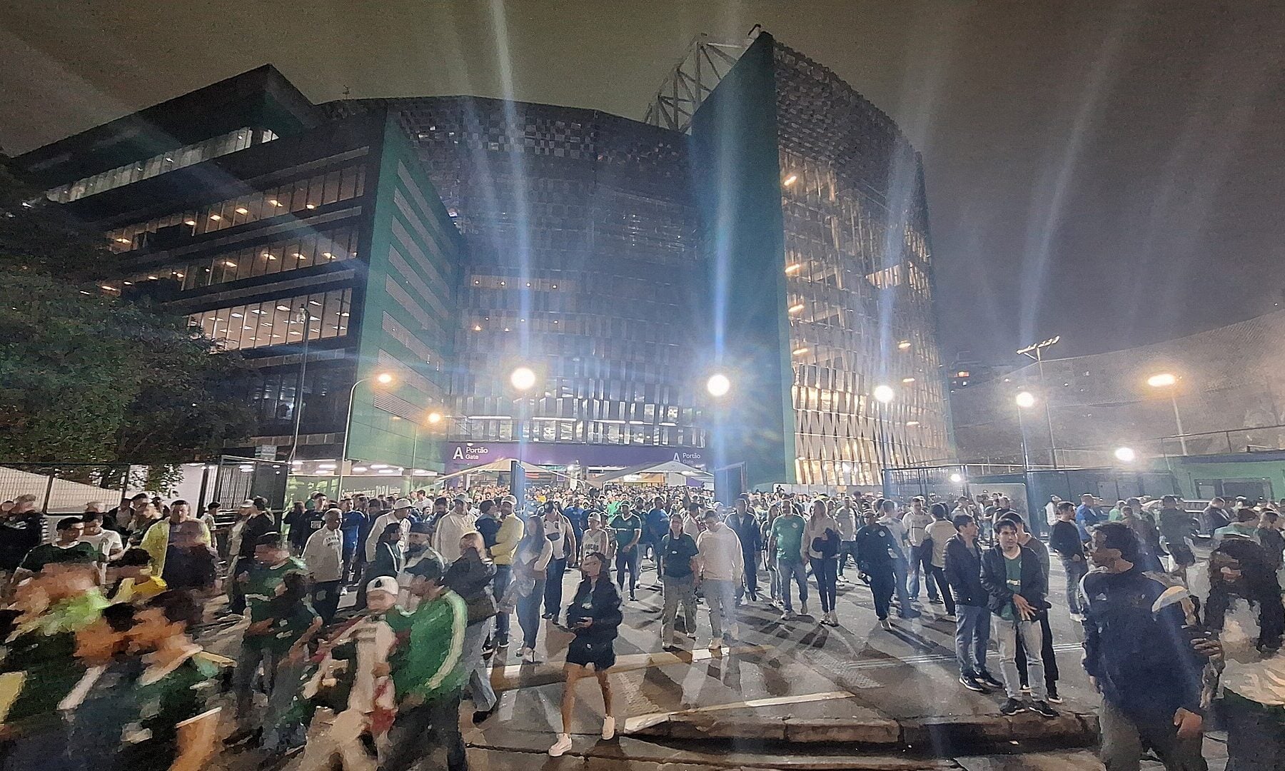Crowds walk across a plaza at night in front of the lit glass and metal facade of Nubank Parque stadium in São Paulo, with tall floodlights and street lamps
