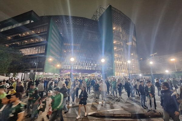 Crowds walk across a plaza at night in front of the lit glass and metal facade of Nubank Parque stadium in São Paulo, with tall floodlights and street lamps