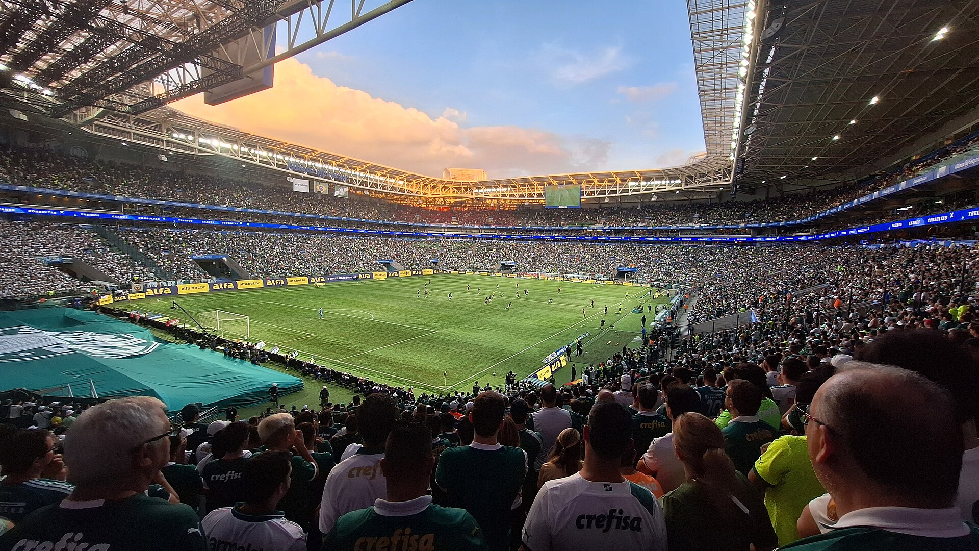 Wide view from the lower stands of a packed stadium in late-afternoon light, with a green football pitch under open sky and a roof over the seating tiers