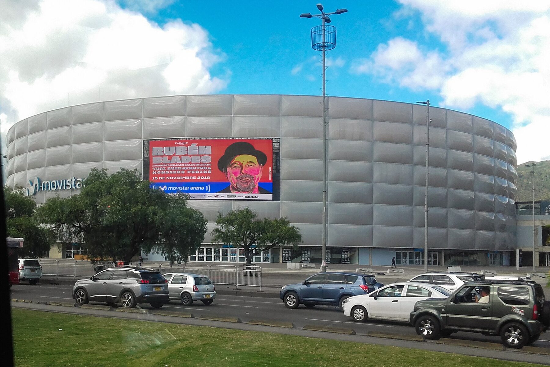 The curved silver facade of Movistar Arena in Bogotá with a large billboard for a Rubén Blades concert, trees and cars passing on the avenue in front