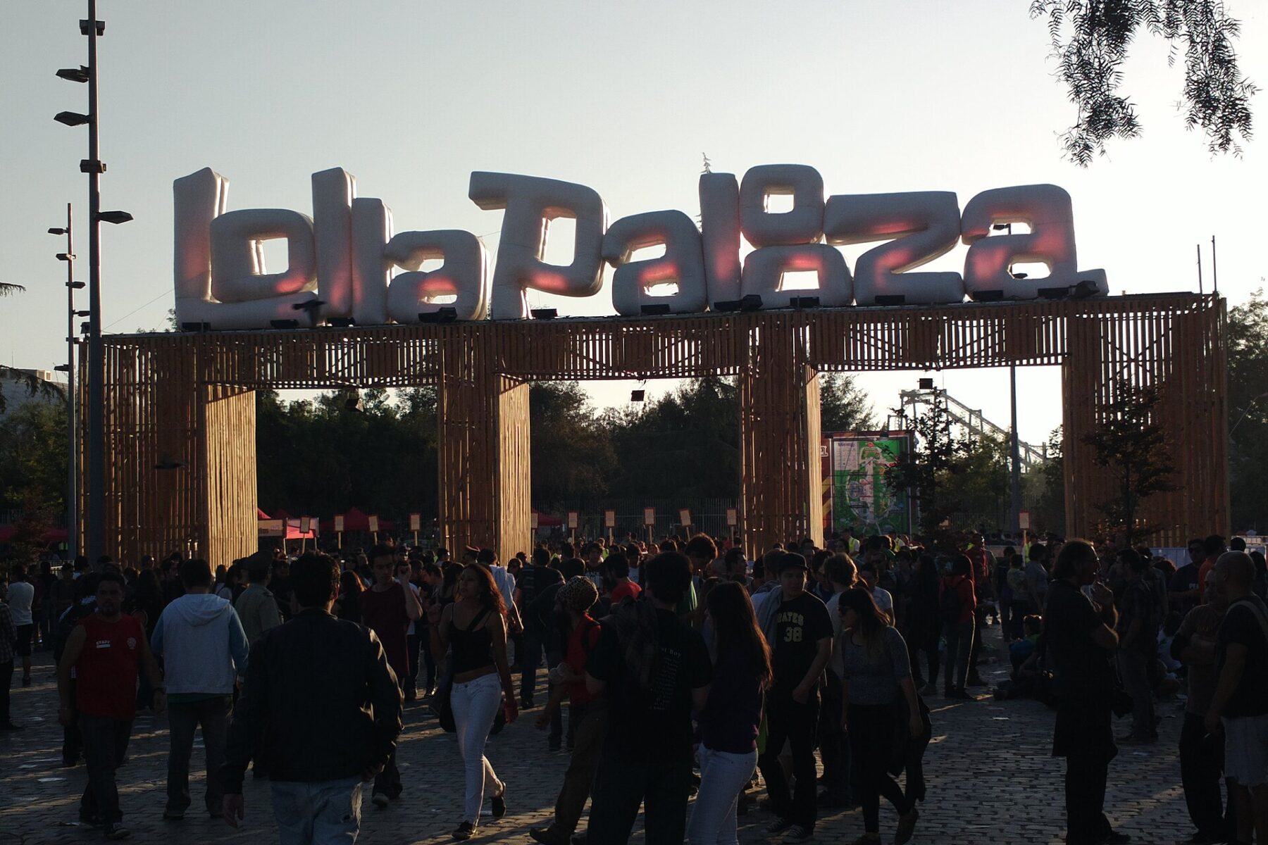 A crowd walks under a wooden entrance gate topped with large inflatable Lollapalooza letters at Parque O'Higgins in Santiago, backlit by low sun