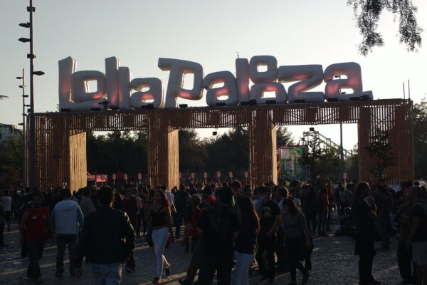 A crowd walks under a wooden entrance gate topped with large inflatable Lollapalooza letters at Parque O'Higgins in Santiago, backlit by low sun