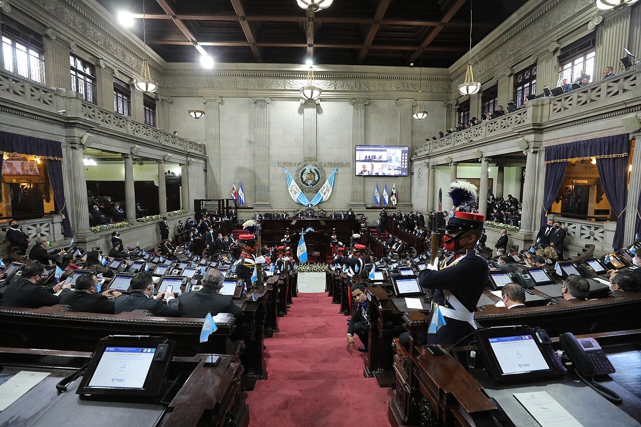 Members of Guatemala's Congress sit at desks in the wood-panelled chamber as honour guards in plumed helmets stand in the aisle