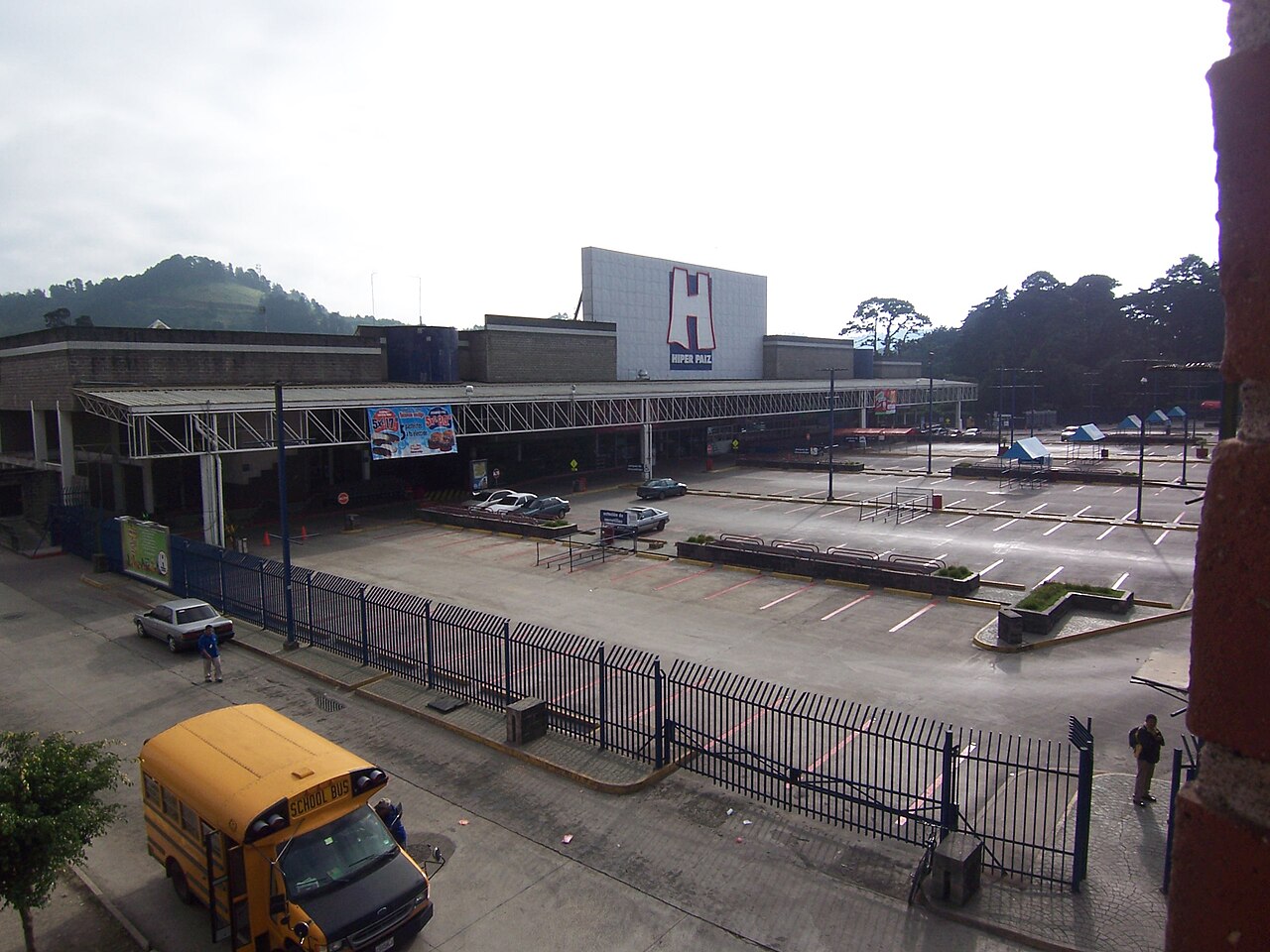 Hiper Paiz hypermarket in Guatemala with a large H sign, a covered entrance, an almost empty car park and a yellow school bus in front