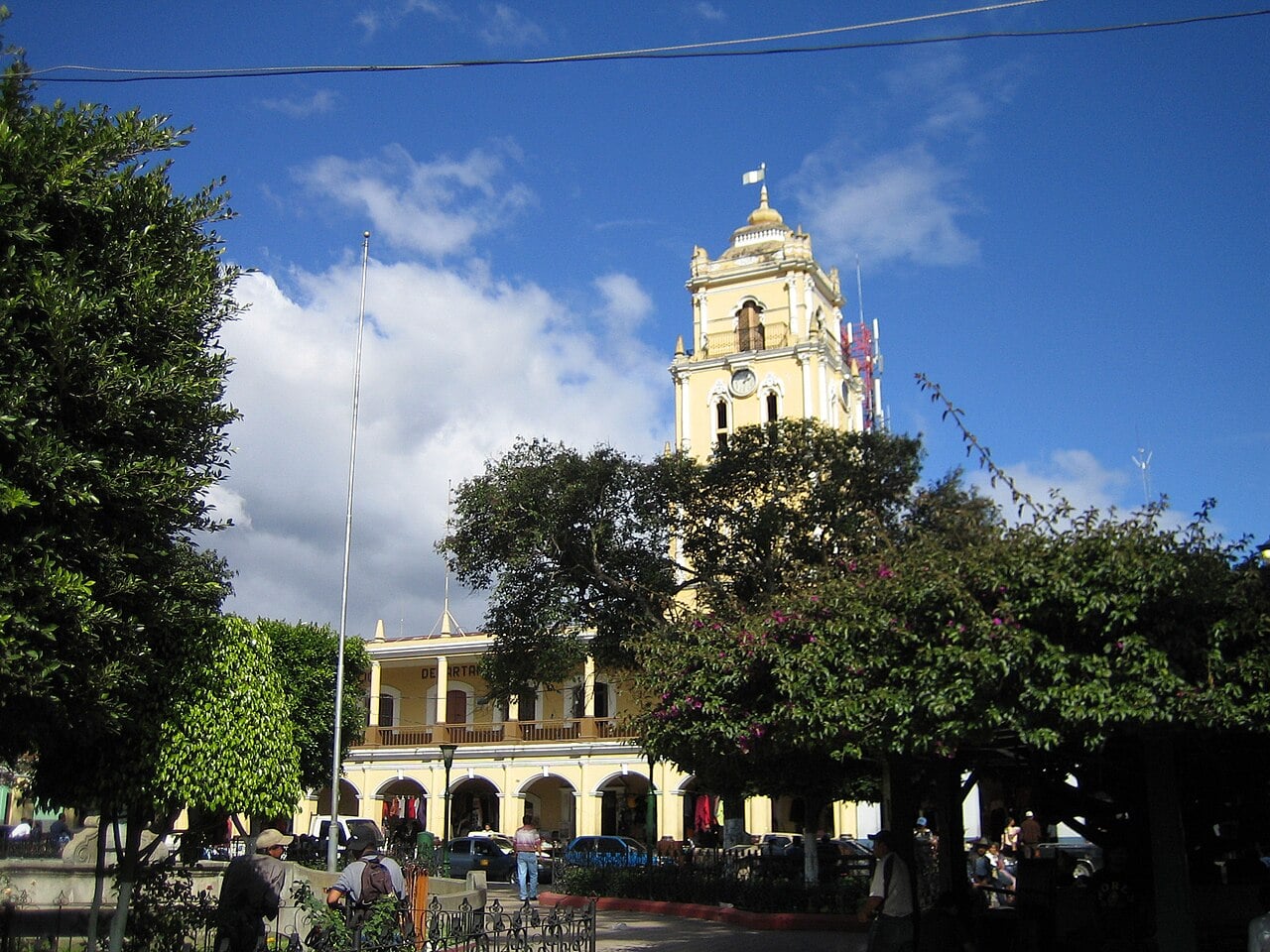 A yellow colonial-style government building with a clock tower behind trees in the central park of Huehuetenango, Guatemala
