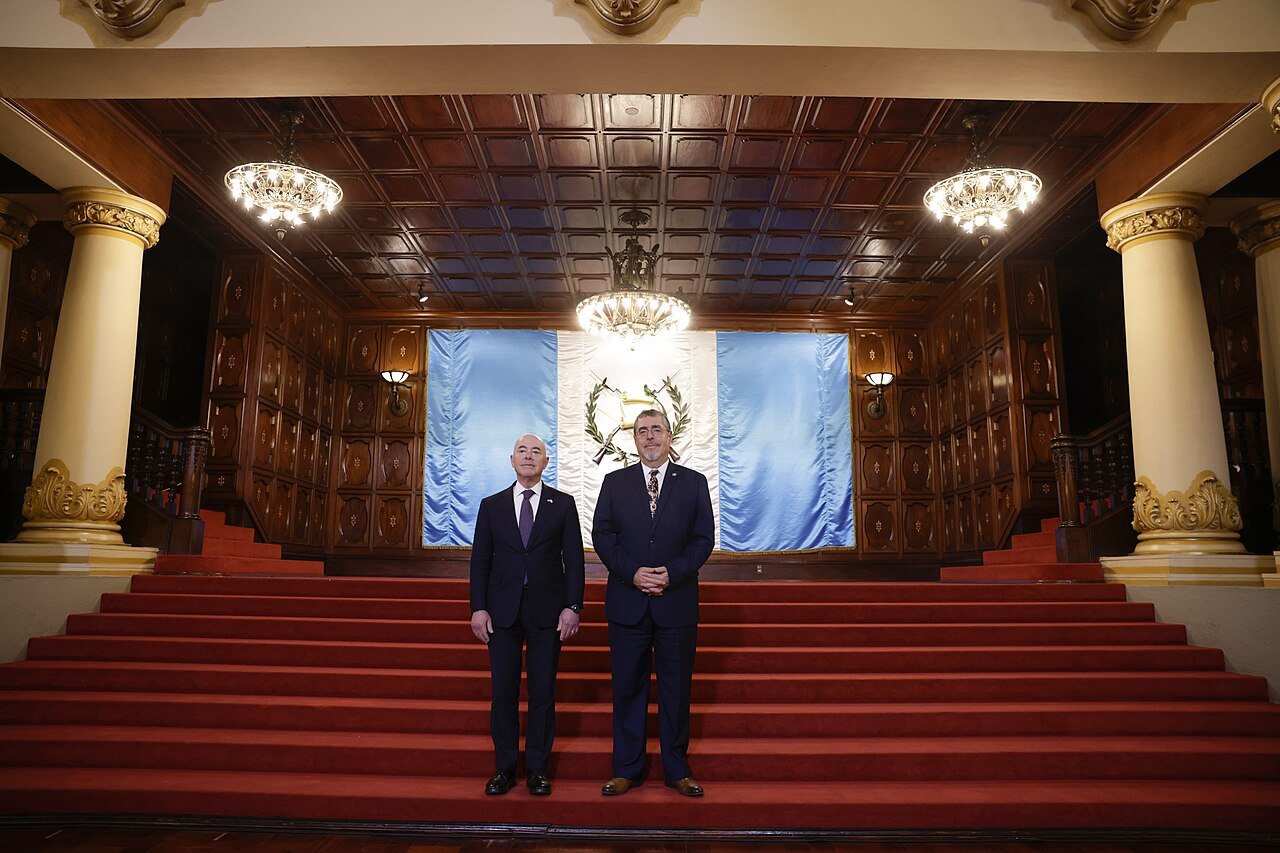 President Bernardo Arévalo standing beside US Homeland Security Secretary Alejandro Mayorkas on red-carpeted steps in front of a Guatemalan flag