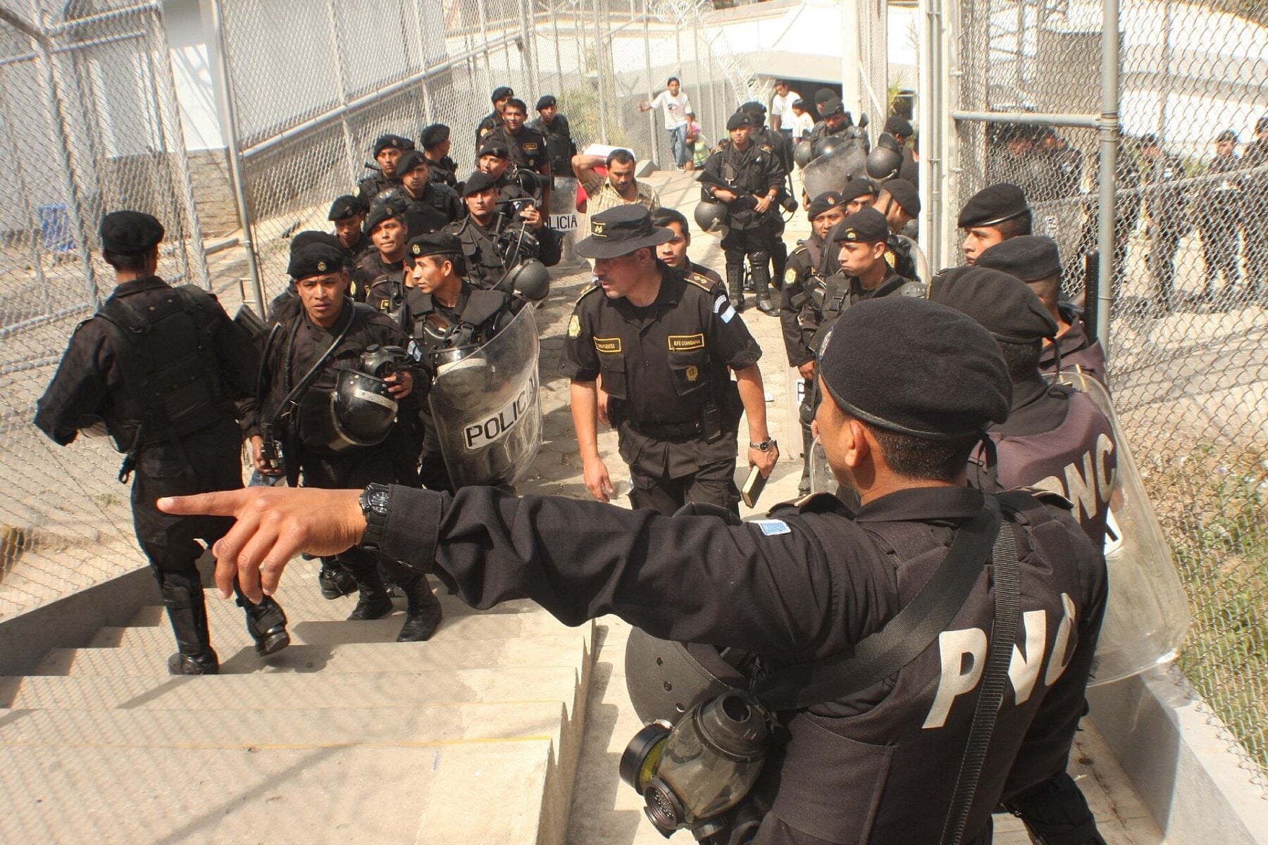 Riot police in black uniforms with helmets and shields climb a concrete stairway between wire fences at a Guatemalan detention centre, while an officer in the foreground points ahead