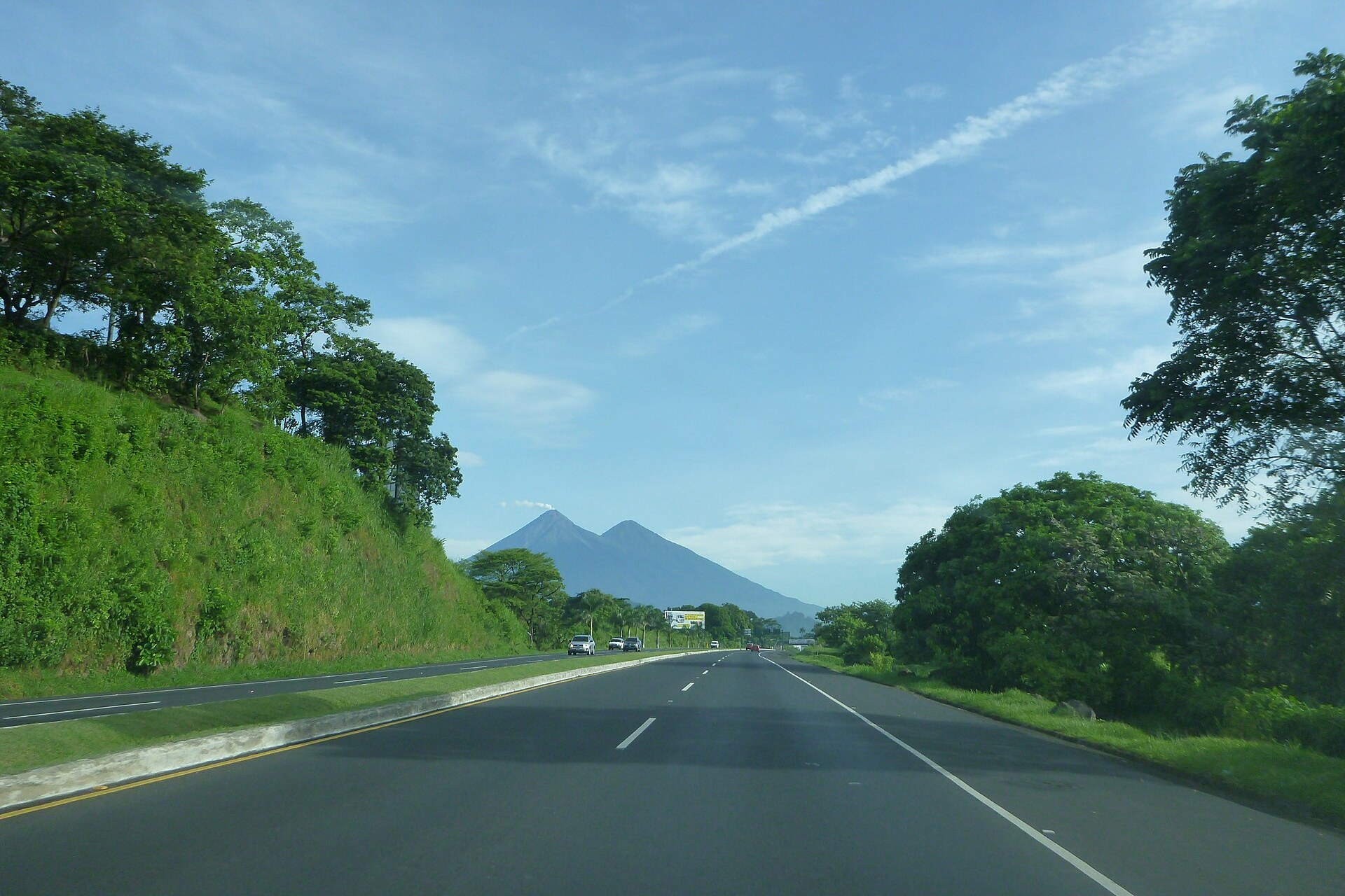 A four-lane highway in Escuintla, Guatemala, with green hillsides on the left and two volcanic peaks on the horizon under a blue sky