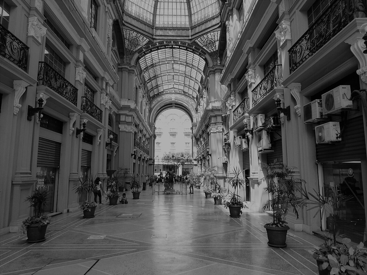 A black-and-white view down a covered arcade in Guayaquil with a glass-vaulted roof, iron balconies and potted plants