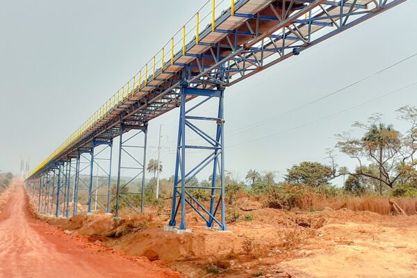 An elevated blue and yellow conveyor gantry runs alongside a red dirt road through green bush in Boffa, Guinea