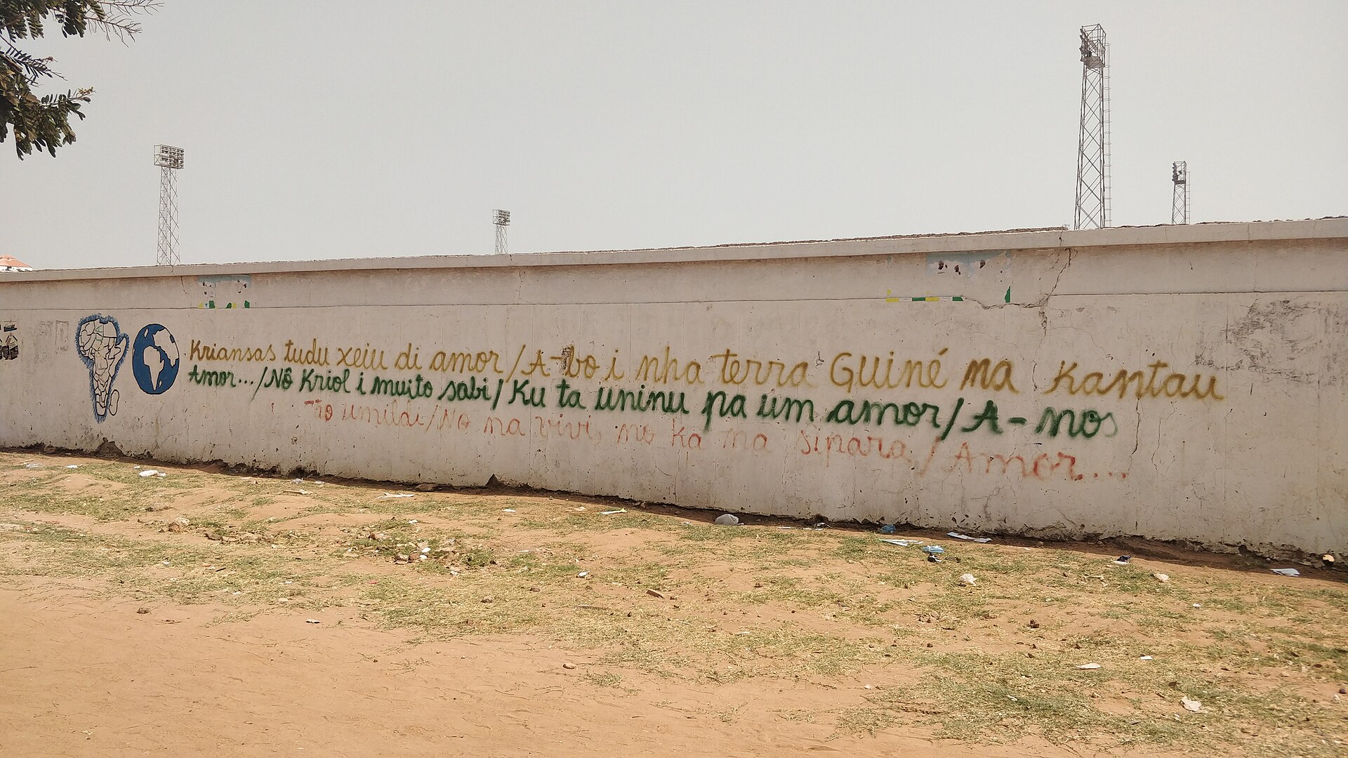 A white wall in Bissau painted with a Crioulo-language message about unity and love, beneath floodlight towers