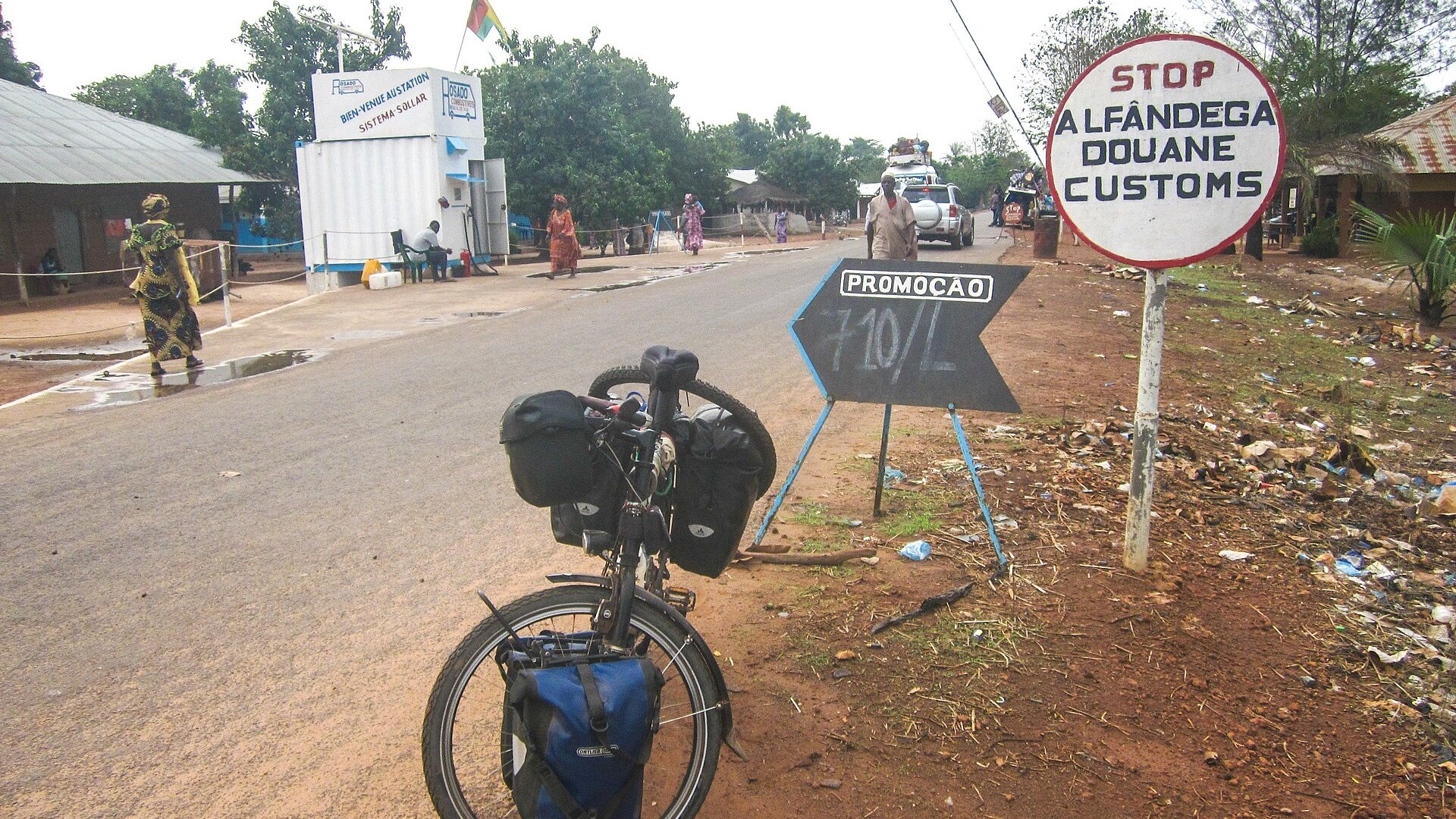 A road at a border crossing between Senegal and Guinea-Bissau, with a customs sign in Portuguese, French and English, a loaded touring bicycle and people walking.