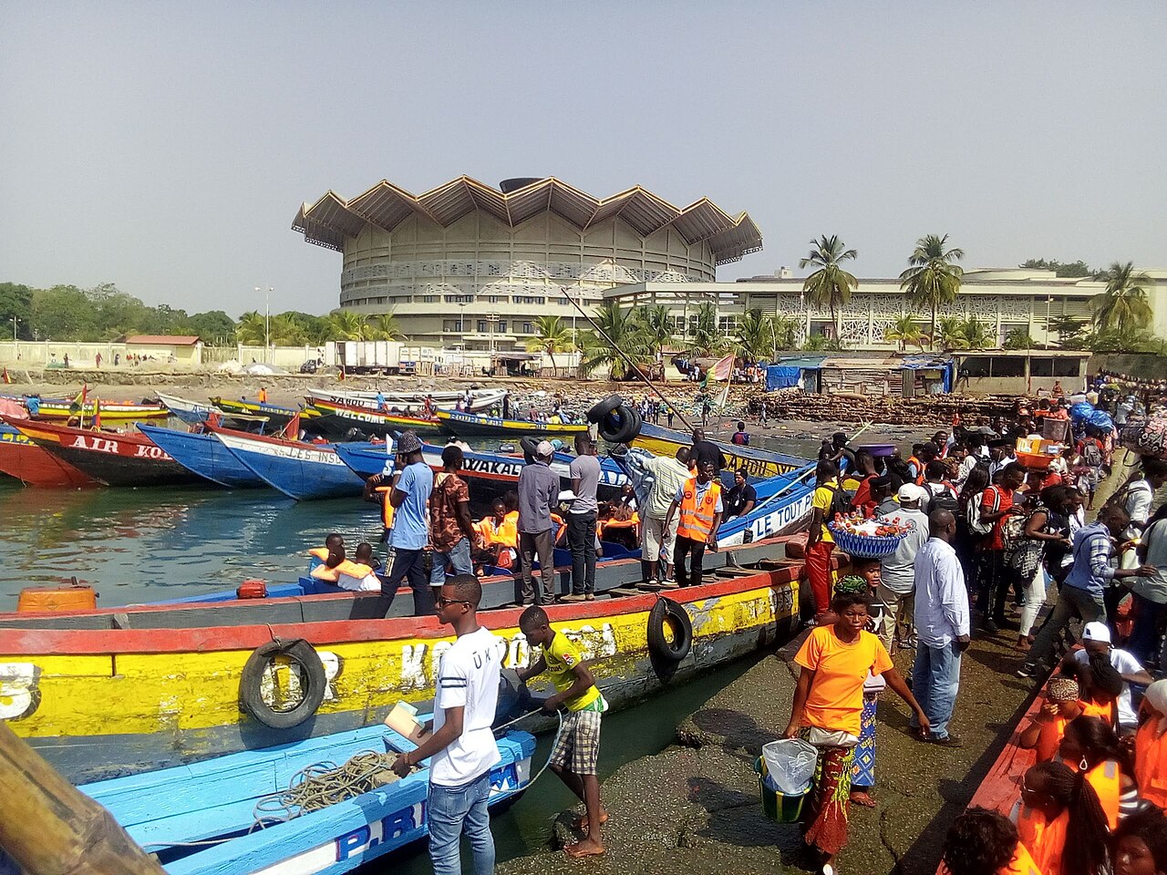 Painted wooden boats and crowds at a landing in Conakry, with a large building with a zigzag roof behind
