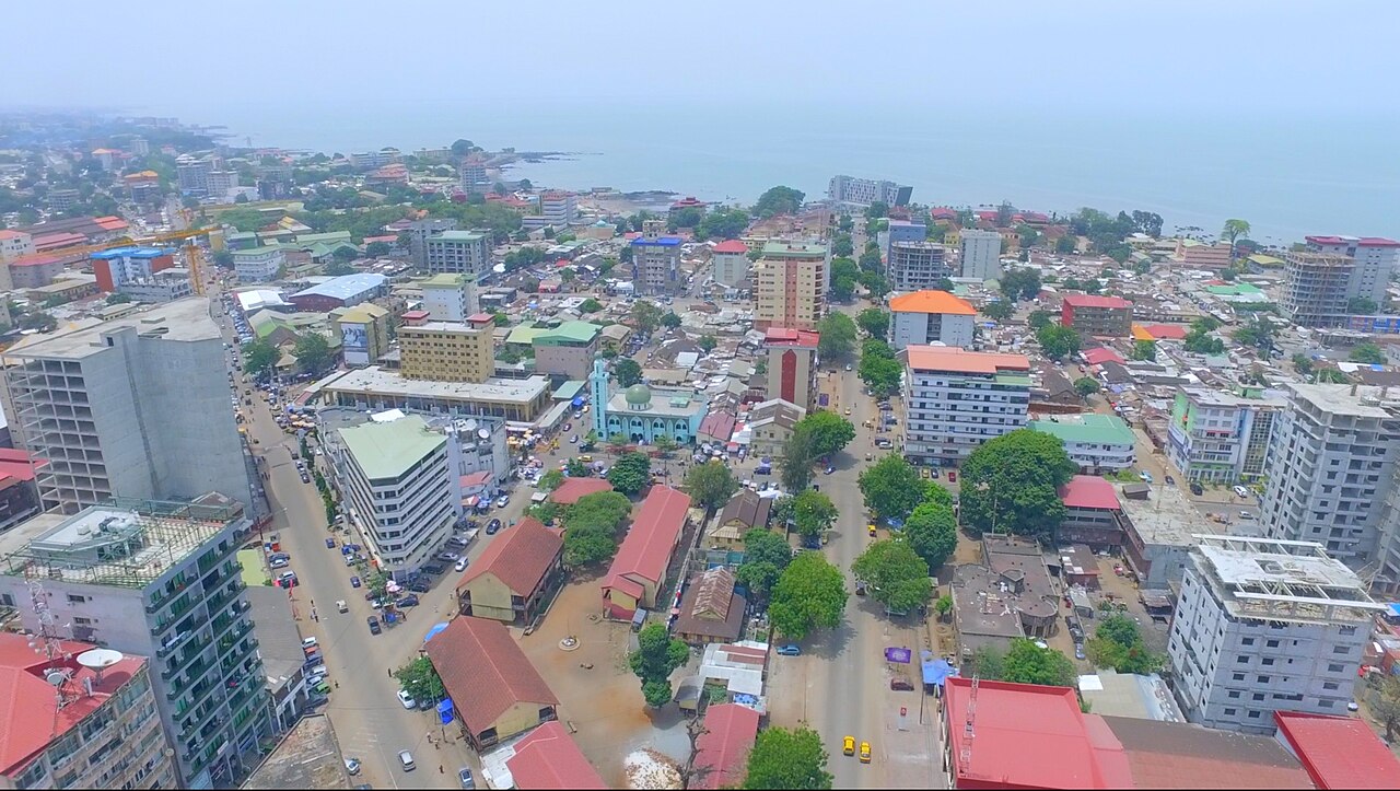 Aerial view of central Conakry with apartment blocks, red roofs, a green-domed mosque and the Atlantic Ocean behind