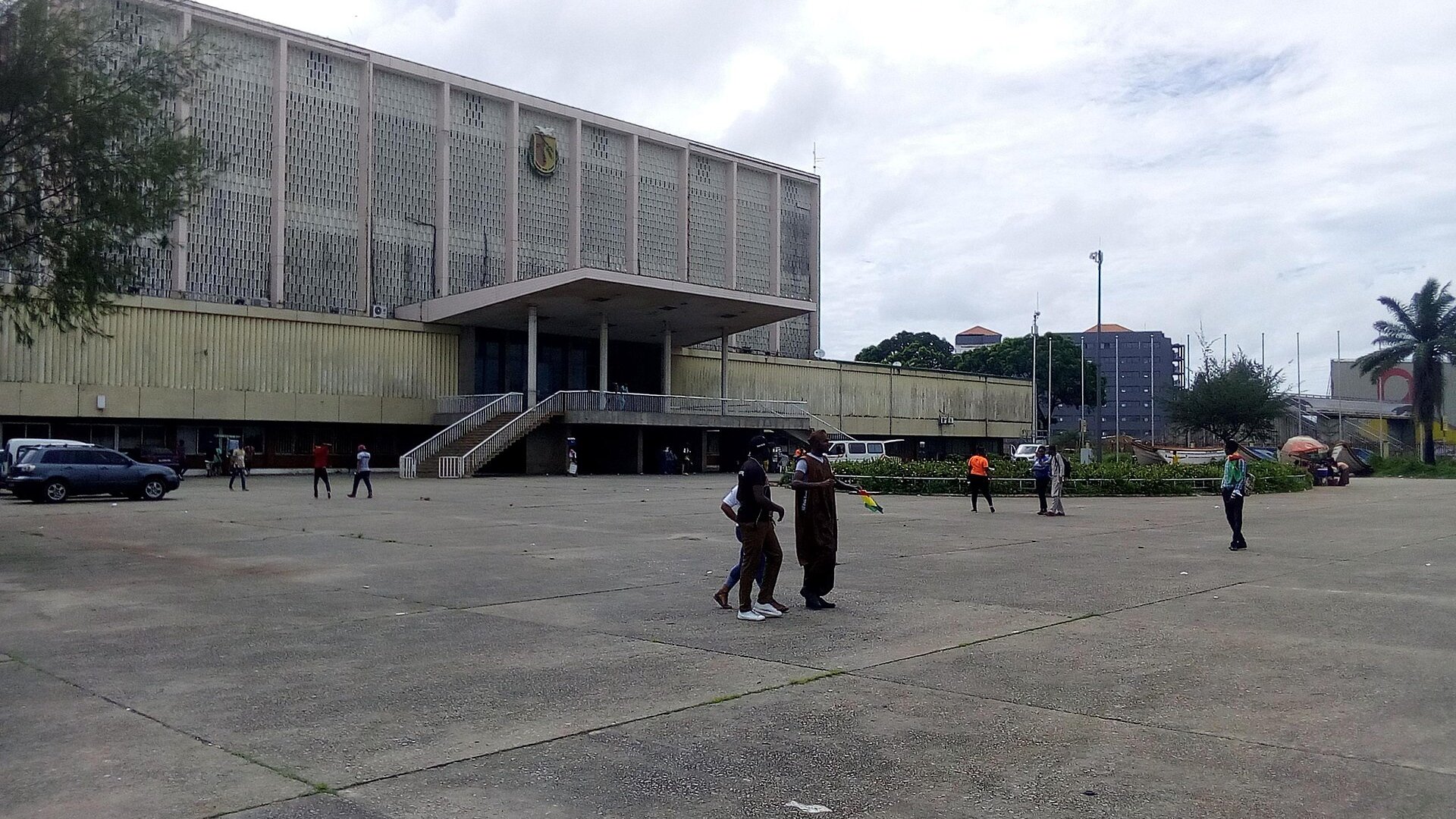 A large mid-century public building with a national emblem on its facade, seen from an open plaza in Conakry, Guinea