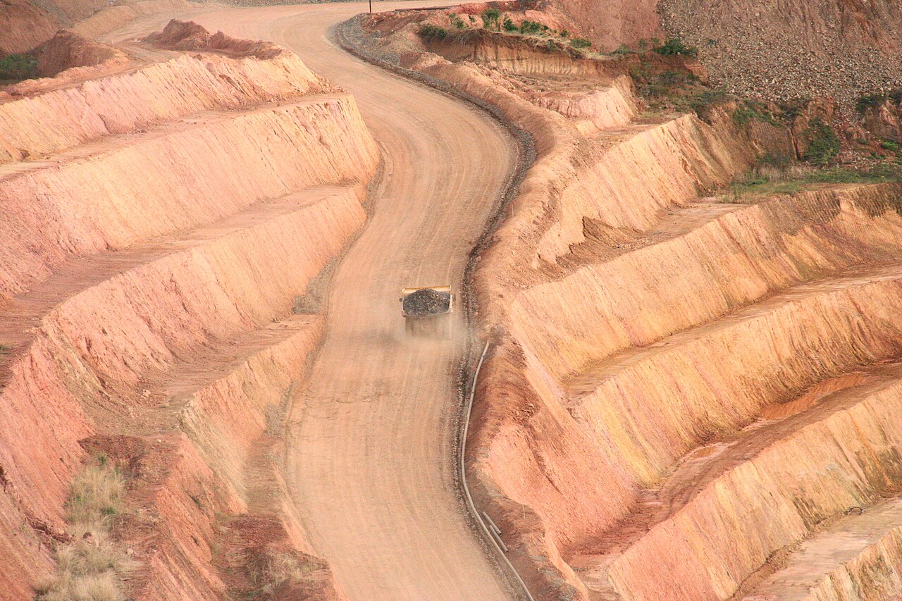 A dump truck loaded with ore climbs a dusty road between terraced red walls of an open-pit gold mine