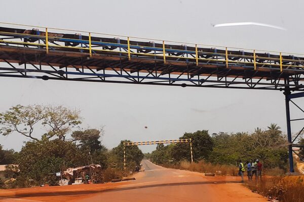 A long overhead conveyor on a steel frame crosses a red dirt road in Boffa, Guinea, with trees, small huts and workers beside the road