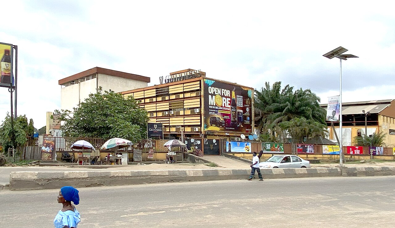 Brick building with a Guinness sign on the roof and a billboard in Lagos, Nigeria, with a pedestrian and parked car in front