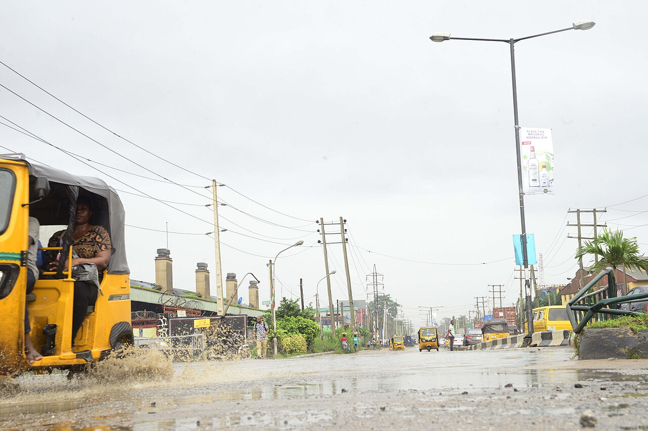 A tricycle taxi splashes through a wet street on Guinness Road in Agege, Lagos, with industrial chimneys behind a fence on the left