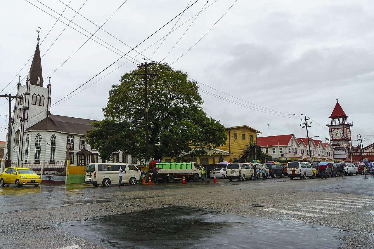 Wet street in central Georgetown with a white wooden church, parked minibuses, a large tree and the red clock tower of Stabroek Market.