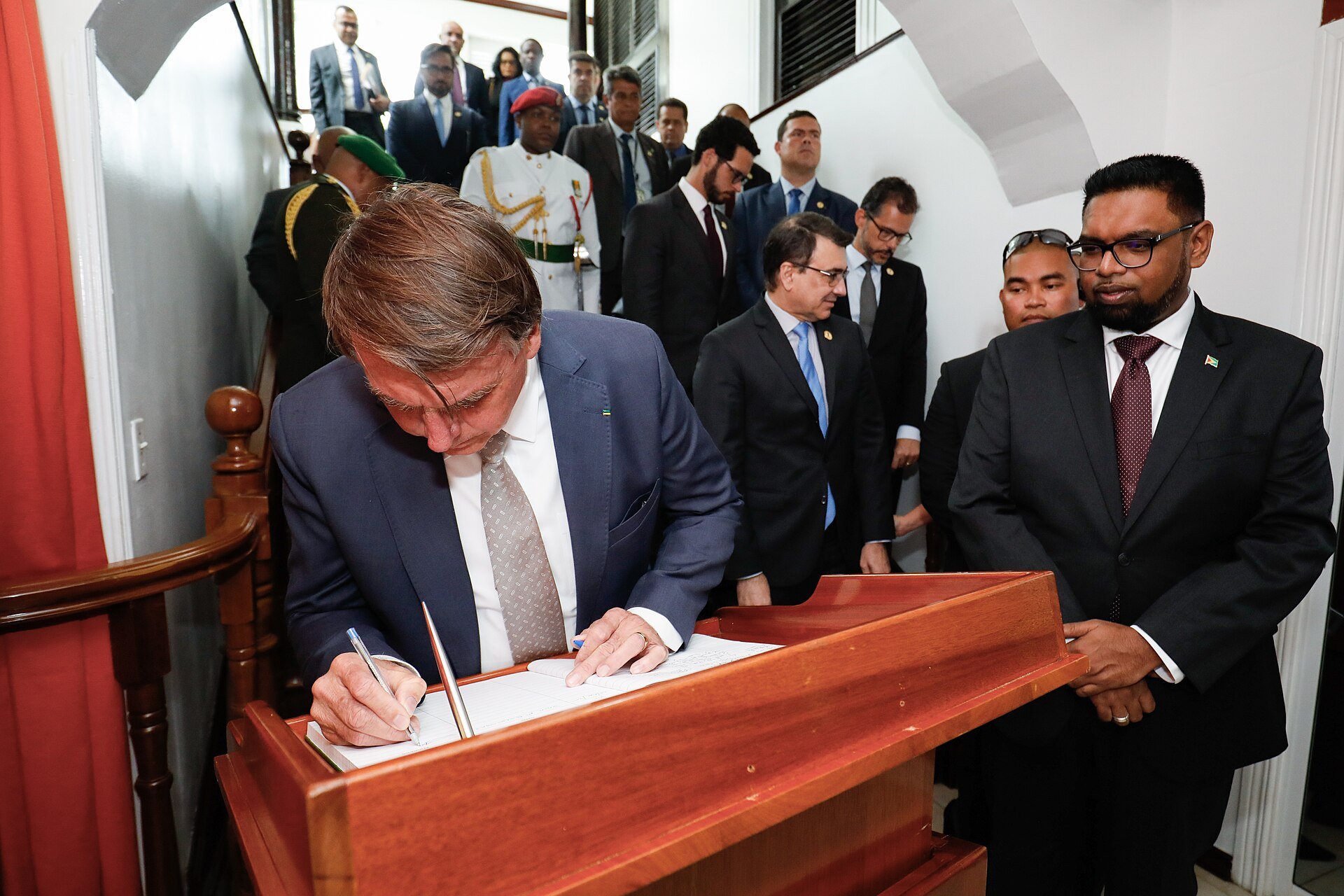Brazil's then president Jair Bolsonaro signing a guest book in Georgetown while Guyana officials look on