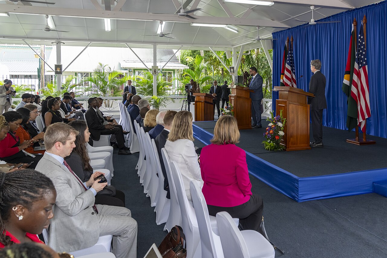 A press event in Georgetown with US and Guyanese flags and two speakers at podiums before an audience