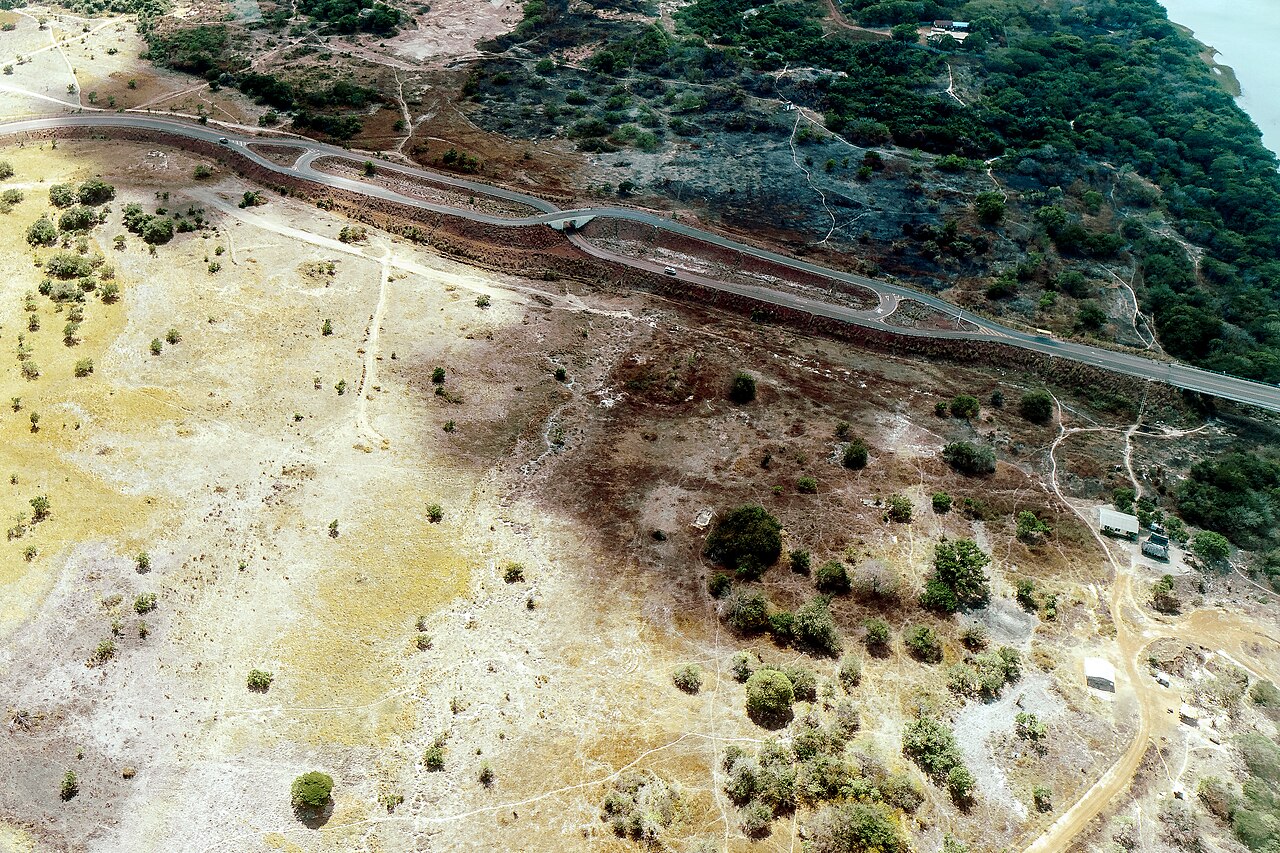 Aerial view of a paved road with a crossover loop running through dry savannah towards a river, with scattered trees and a few buildings