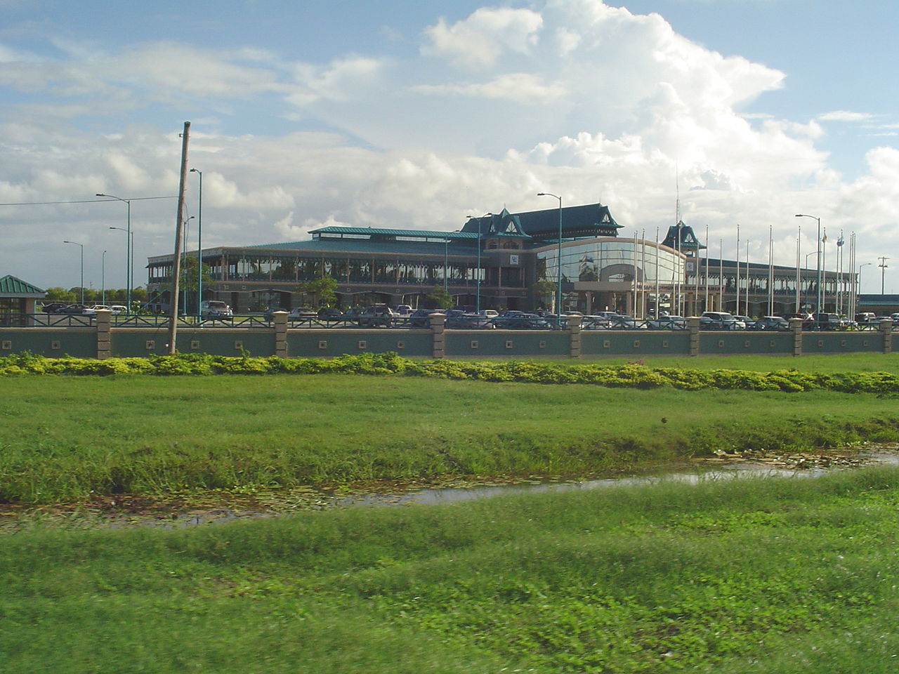 A long two-storey building with green pagoda-style roofs, a glass entrance and a row of flagpoles behind a wall and a grass verge