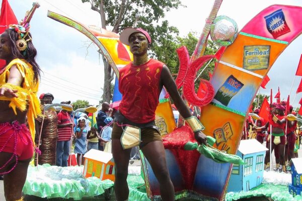 A costumed performer in red and gold dances beside a colourful float in a Mashramani parade in Guyana