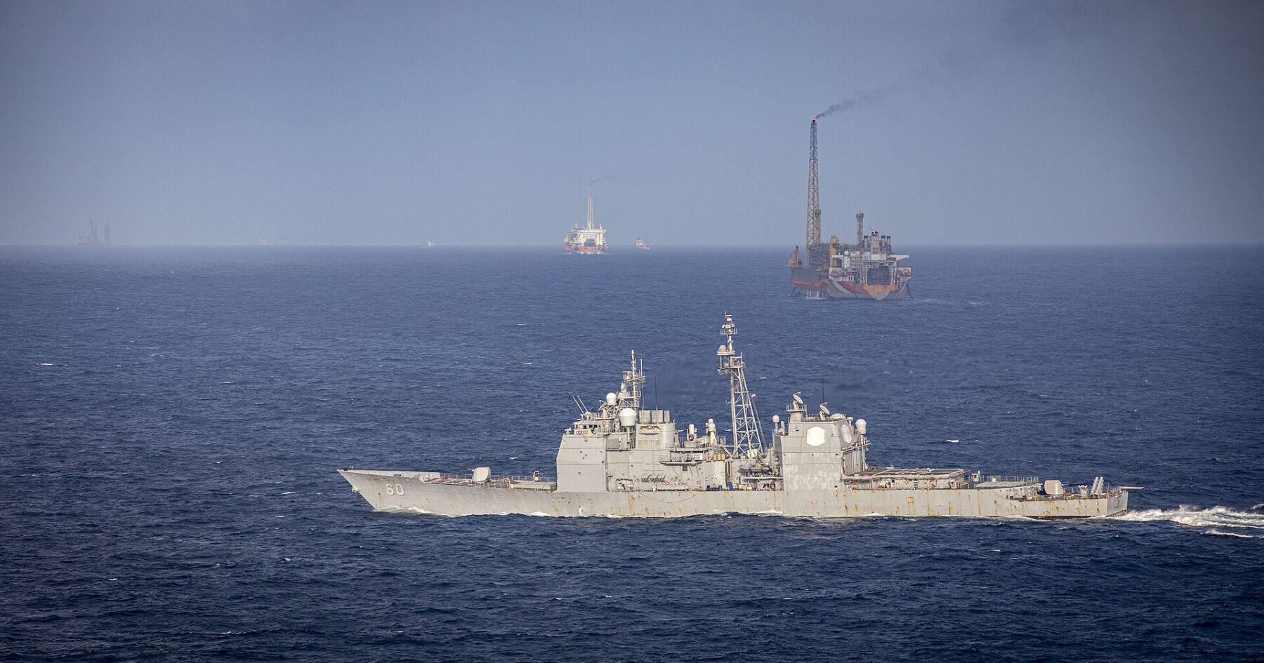 A US Navy cruiser passes a floating production vessel with a flare in the Stabroek Block off Guyana