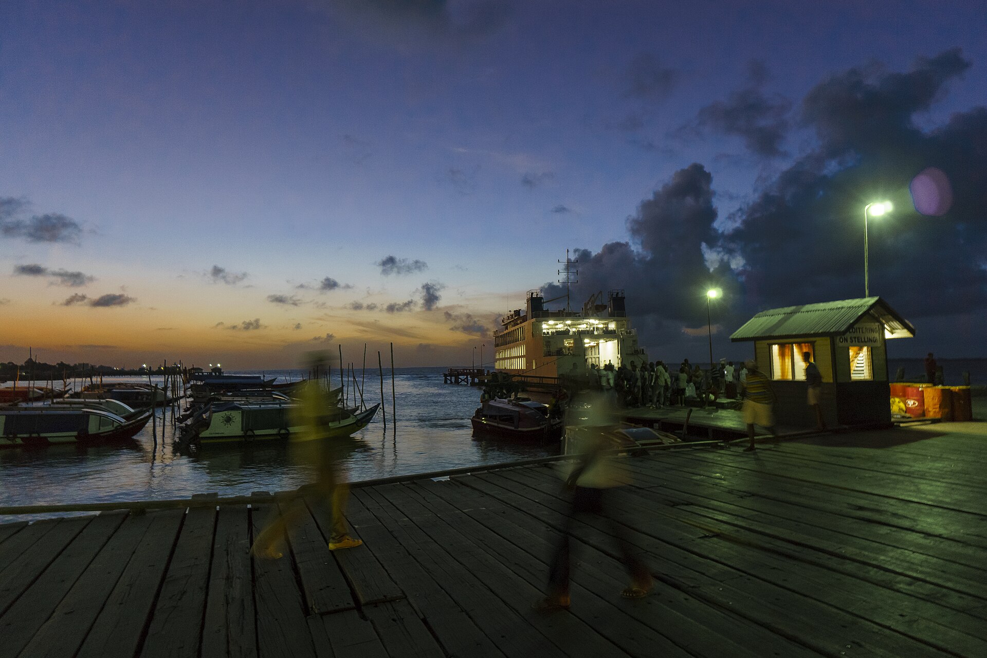 A large passenger ferry moored at a wooden ferry stelling at dusk in Guyana, with small boats alongside