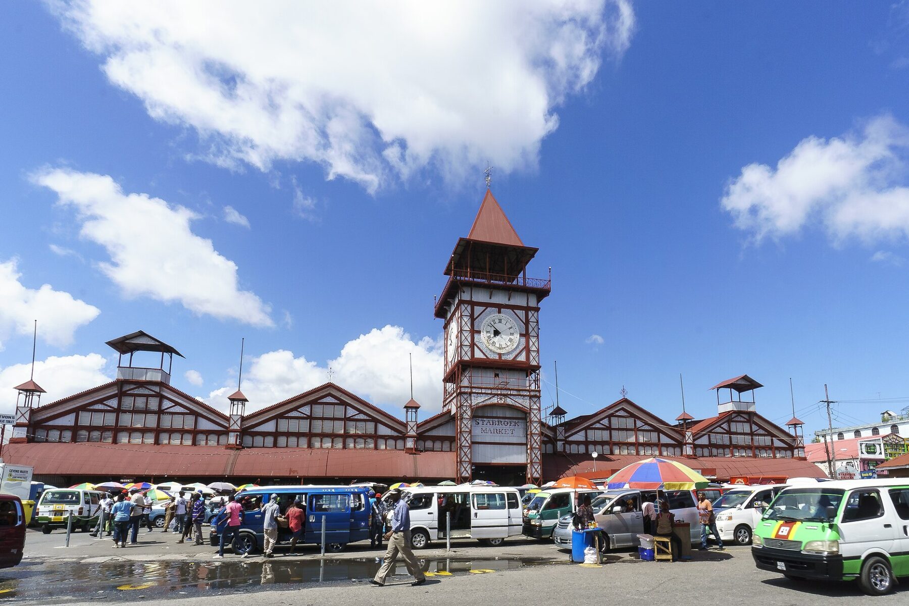Stabroek Market's red clock tower and iron market hall in Georgetown, Guyana, with minibuses, umbrellas of street vendors and shoppers in front under a blue sky