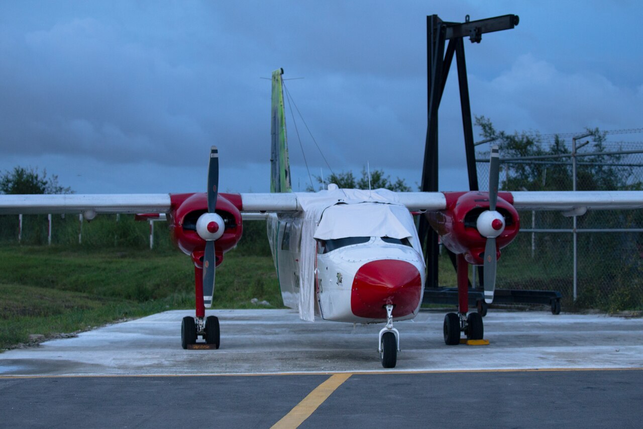 A white and red twin-propeller light aircraft with a covered windscreen parked on an apron under a cloudy sky