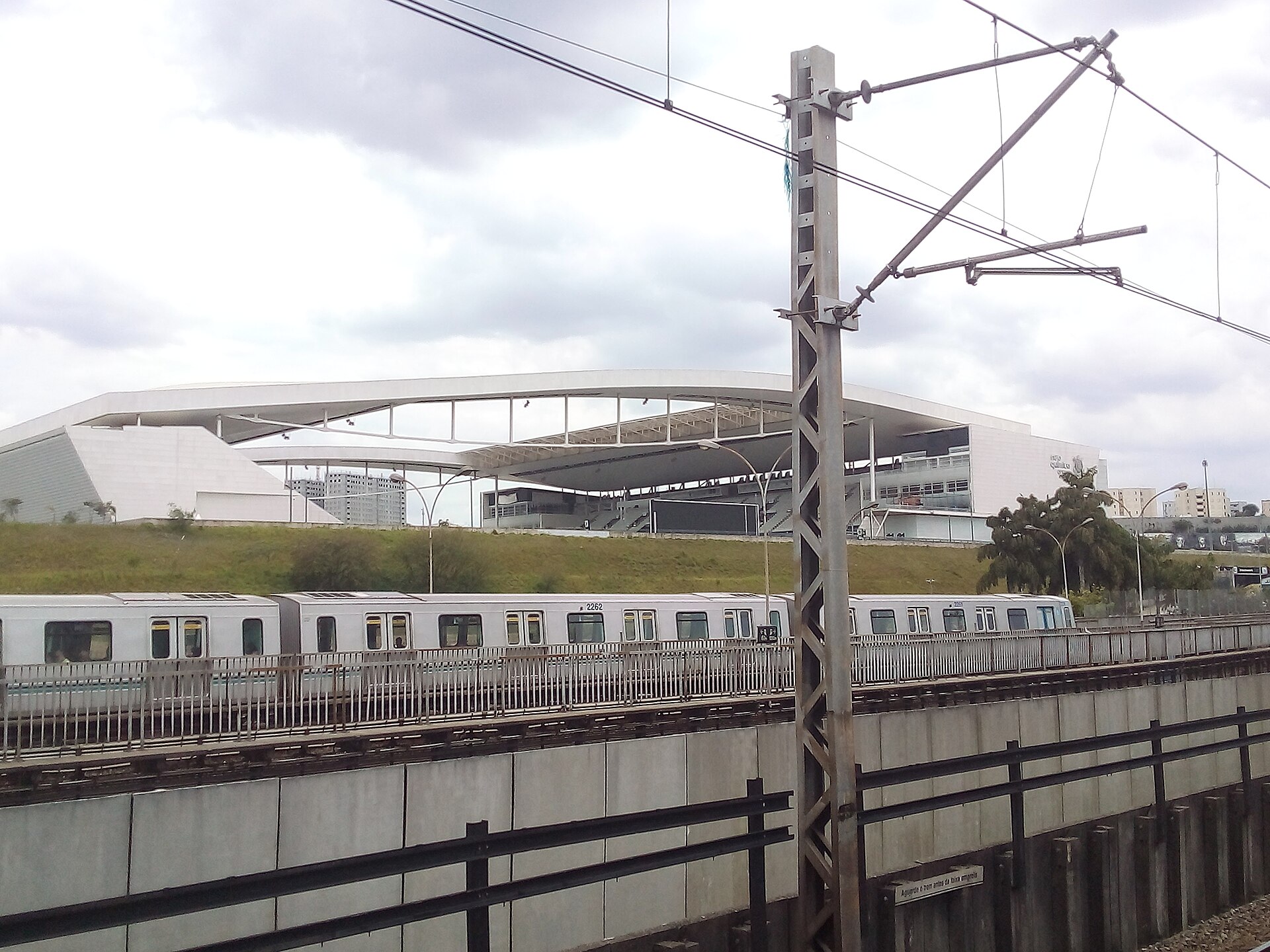 Neo Química Arena's white roof rim behind a grass embankment, with a metro or commuter train on elevated tracks in the foreground