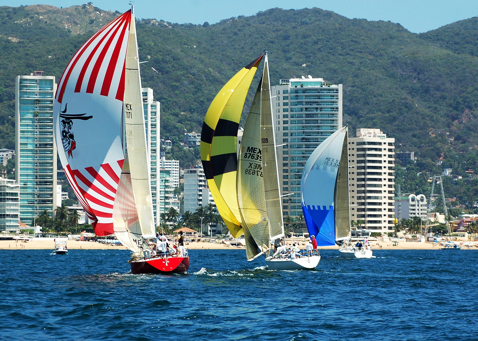 Racing sailboats with red-and-white, yellow-and-black and blue spinnakers off a beach lined with high-rise hotels and green hills