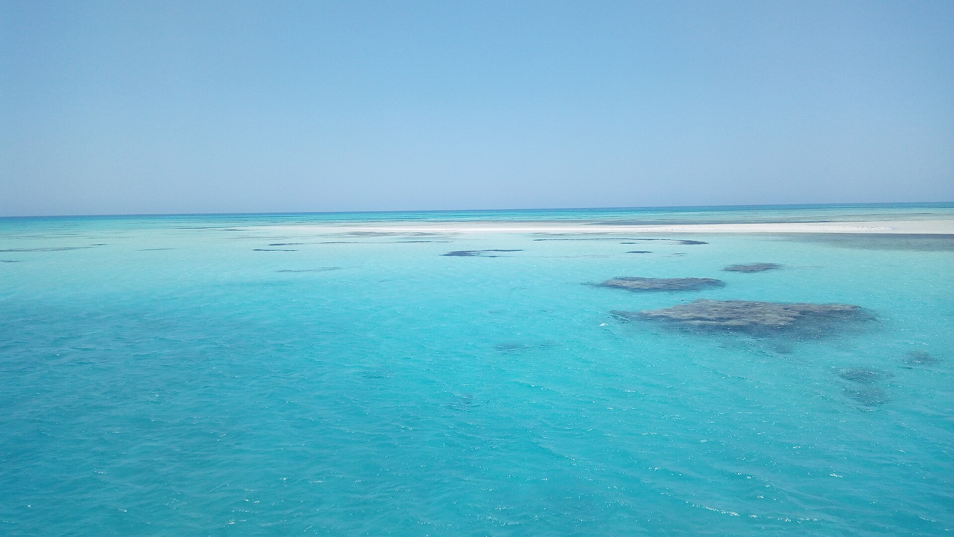 Shallow turquoise Red Sea water with dark coral patches and a white sandbar on the horizon